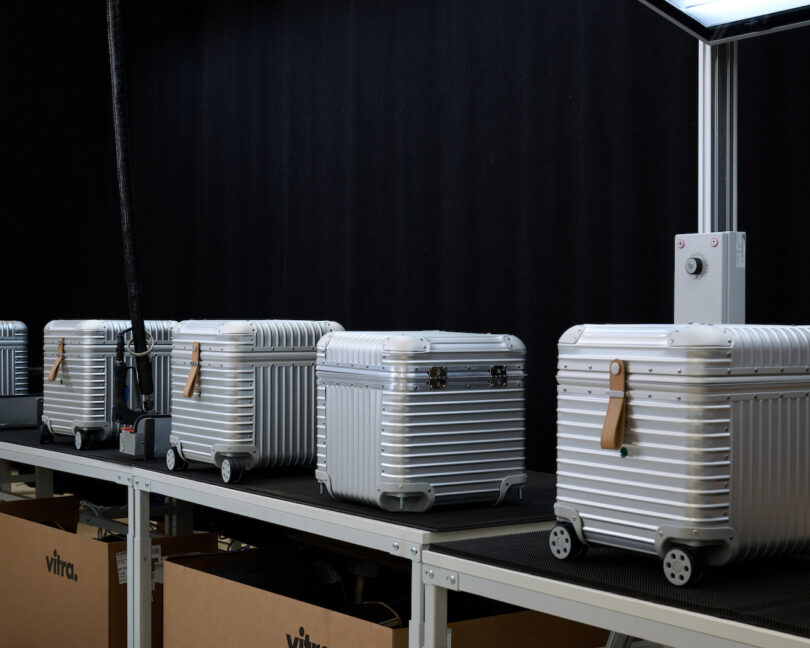 Four silver hard-shell suitcases with brown tags are lined up on a conveyor belt under a light, against a black background