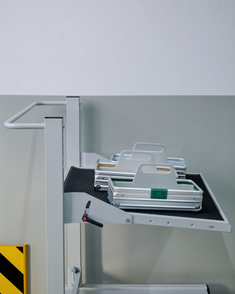 A metal cart with a slanted shelf holds two aluminum carrying cases with handles, positioned against a two-tone wall with a yellow and black striped floor sign nearby