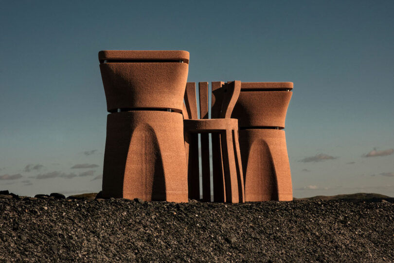 Three large, sculptural, rust-colored structures stand on a barren, rocky landscape under a clear sky
