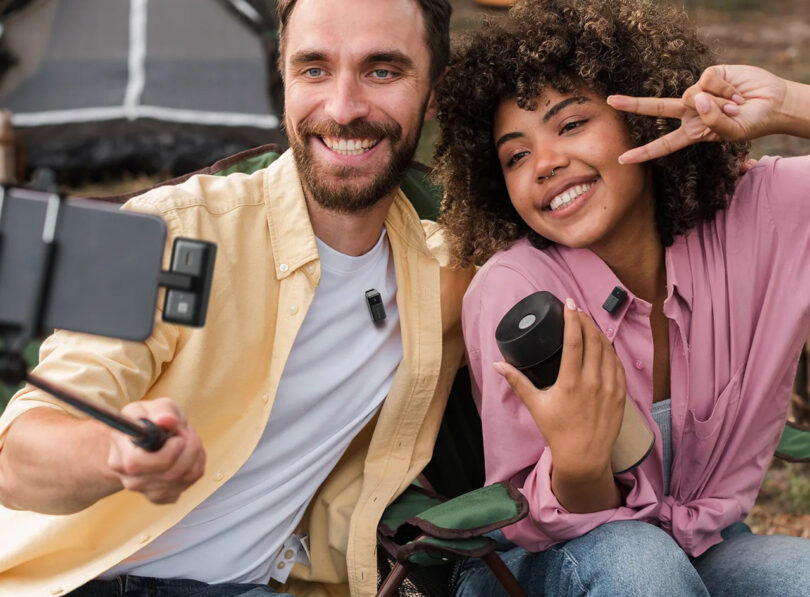 Two people sitting outdoors, smiling and taking a selfie with a Saramonic Air-equipped smartphone on a selfie stick. One holds a drink and makes a peace sign as camping gear is visible in the background.