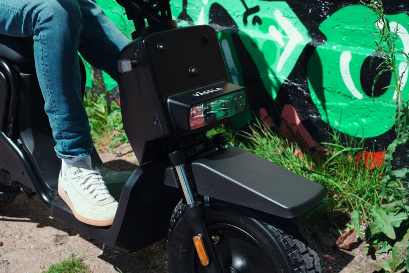 A person wearing white sneakers sits on a black Virta electric scooter parked beside a colorful graffiti wall.