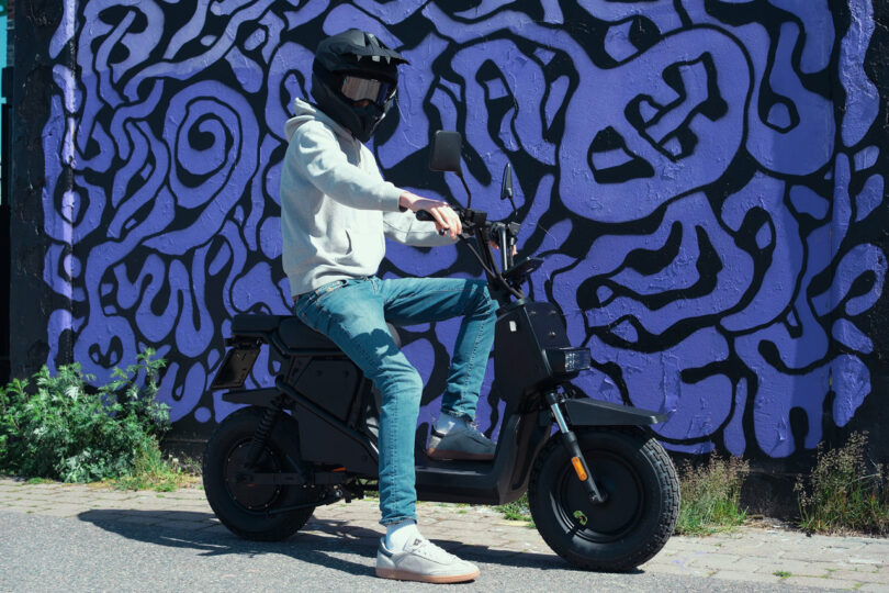 A person wearing a helmet and hoodie sits on a black electric scooter in front of a purple and black abstract mural.