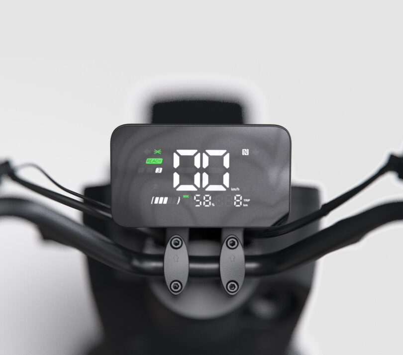 Close-up of a digital display on a motorcycle handlebar showing speed, gear, battery level, and other indicators against a plain background.