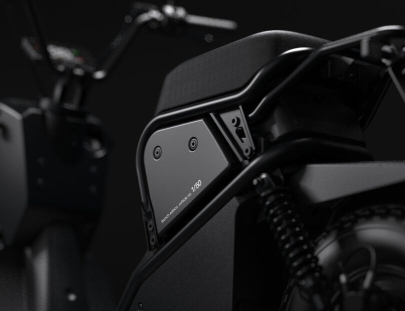 Close-up of a matte black electric motorcycle focusing on the frame, battery compartment, and rear suspension with blurred handlebars in the background.