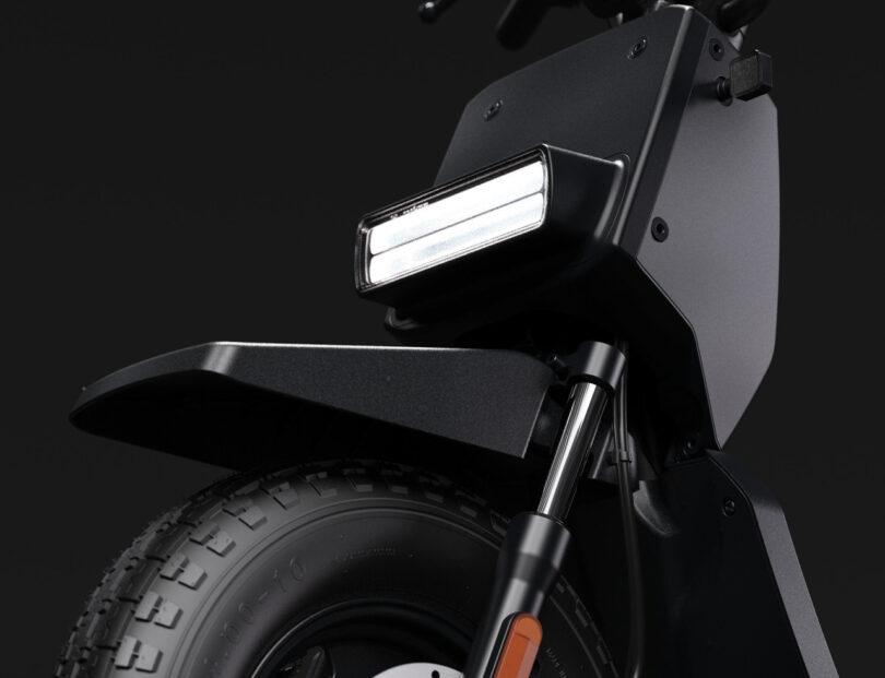 Close-up of the front section of a black electric scooter, showing the headlight, suspension fork, and part of the tire against a dark background.
