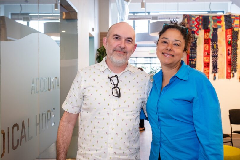 A man and a woman stand together and smile in an office lobby with glass doors and colorful fabric art on the wall in the background