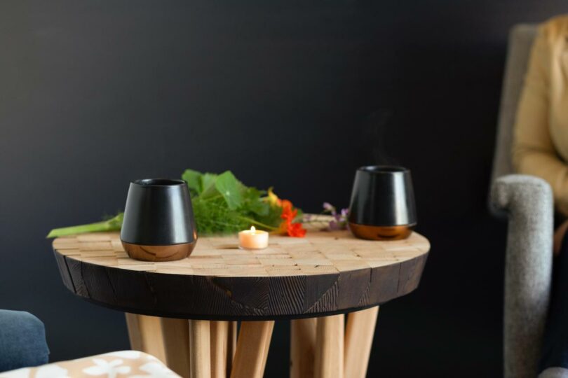 A round wooden table with two black cups, a small lit candle, and a green and orange floral arrangement; part of a person is seated on a gray chair nearby