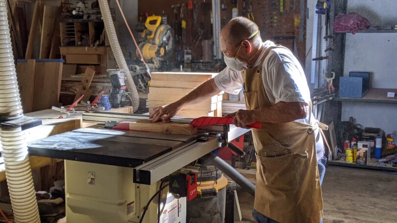 A man wearing safety glasses and a mask operates a table saw to cut a piece of wood in a workshop