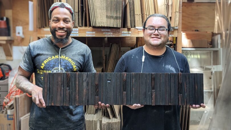 Two men standing in a workshop hold a rectangular wooden panel made of vertically aligned dark wood pieces; shelves with materials are visible in the background