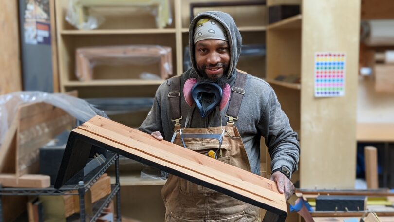 A person in overalls and a hoodie holds a wooden project in a workshop, with shelves and various tools visible in the background