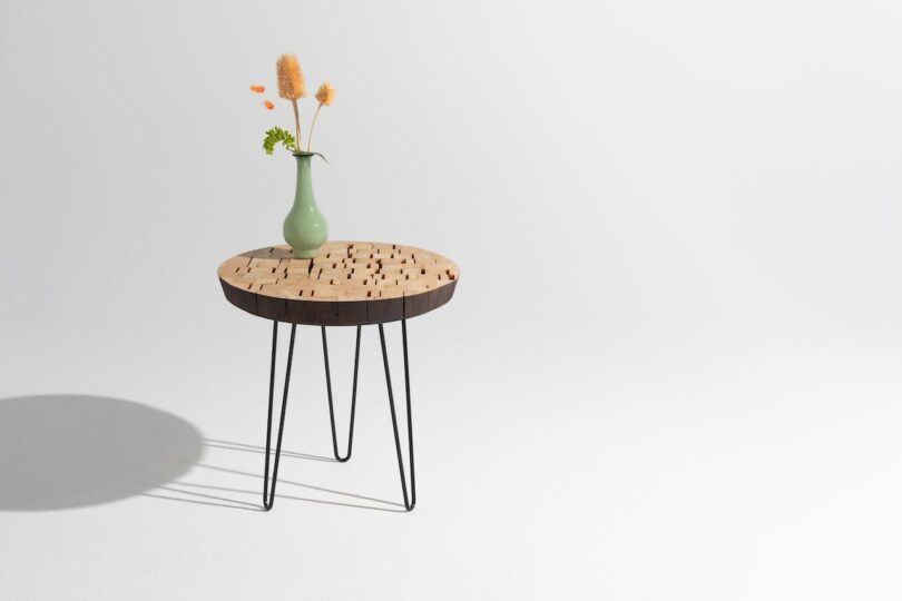 A round wooden table with black metal hairpin legs holds a green vase with dried flowers, set against a plain white background