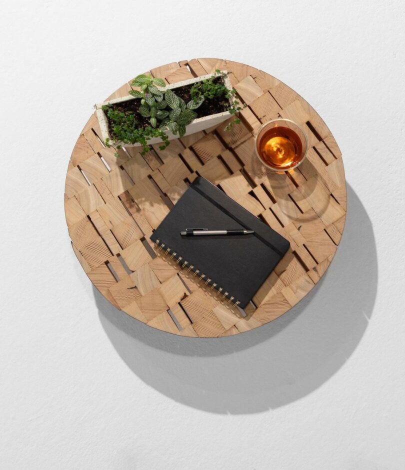 A round woven wooden table with a black notebook, a pen, a small rectangular plant pot, and a glass of amber liquid on top