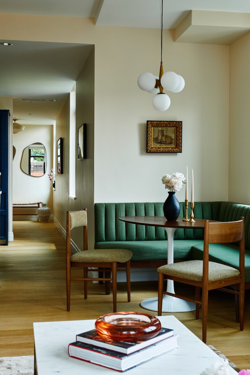 A modern dining nook with a green upholstered banquette, round table, two wooden chairs, a vase with flowers, and a pendant light. A hallway with mirrors is visible in the background.