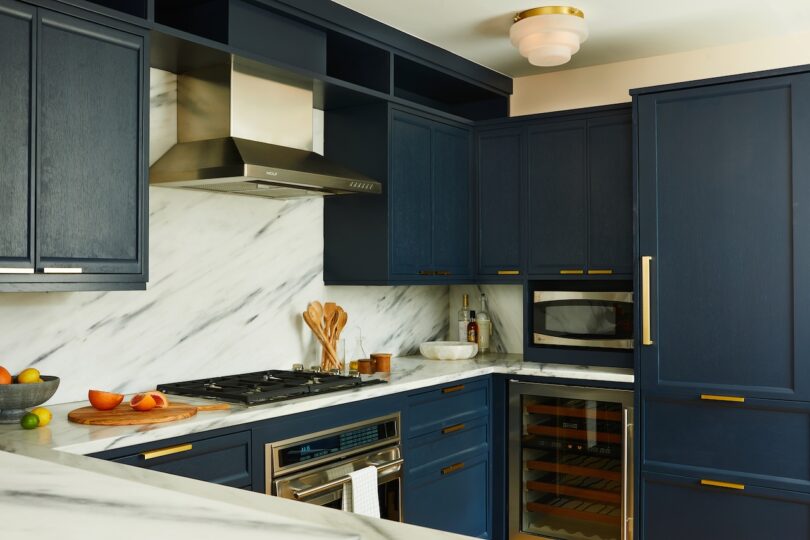 Modern kitchen with navy blue cabinets, marble countertops and backsplash, stainless steel appliances, and gold hardware. Fruits and utensils are on the counter.