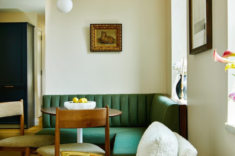 A dining nook with a green upholstered bench, a round wooden table, a bowl of lemons, and framed art on cream-colored walls.