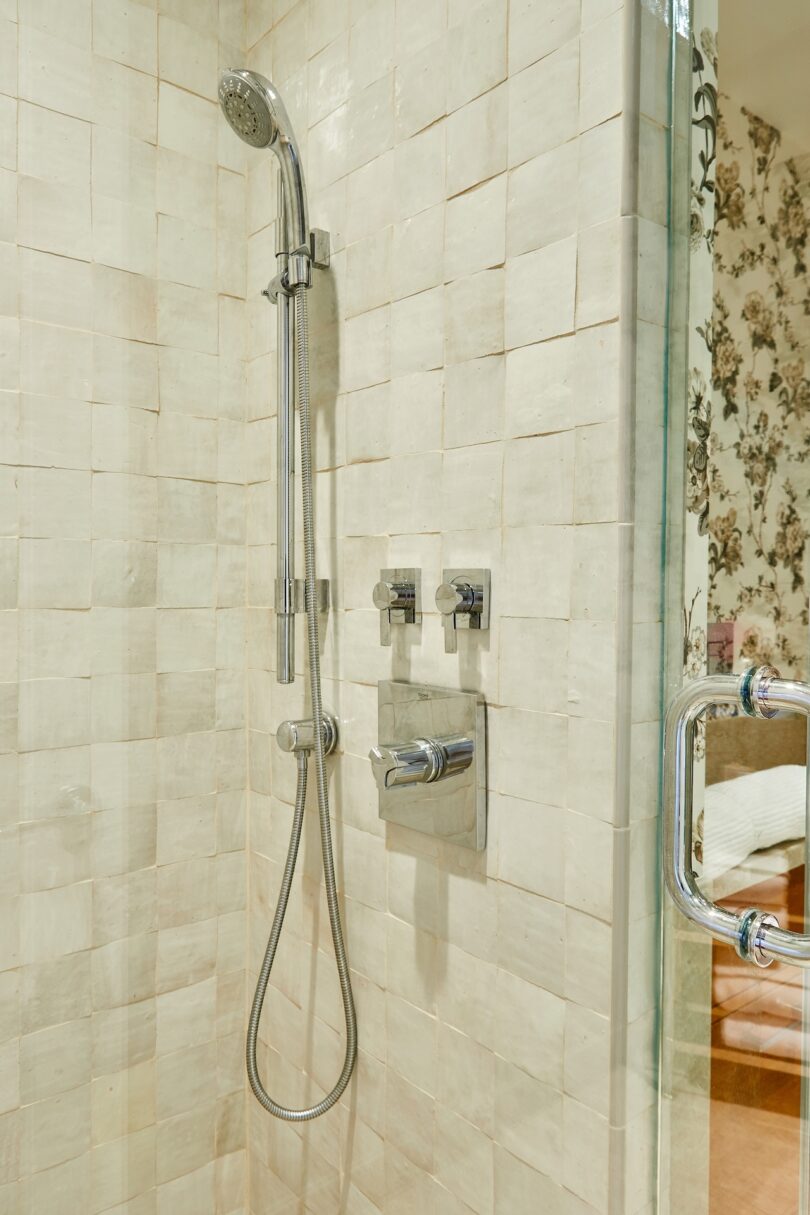 Modern shower with a handheld showerhead, chrome fixtures, and square beige tiles. Glass shower door partially visible on the right. Floral wallpaper can be seen outside the shower.