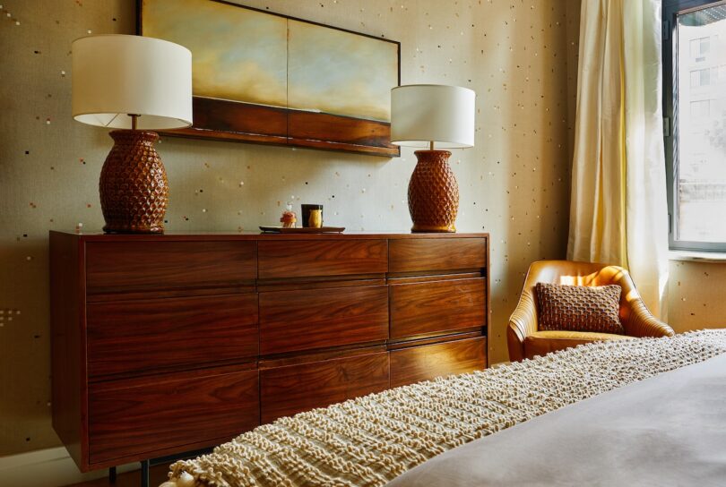 A bedroom with a wooden dresser, two matching lamps, a painting above, a yellow armchair with a pillow, beige walls, and natural light from a window.