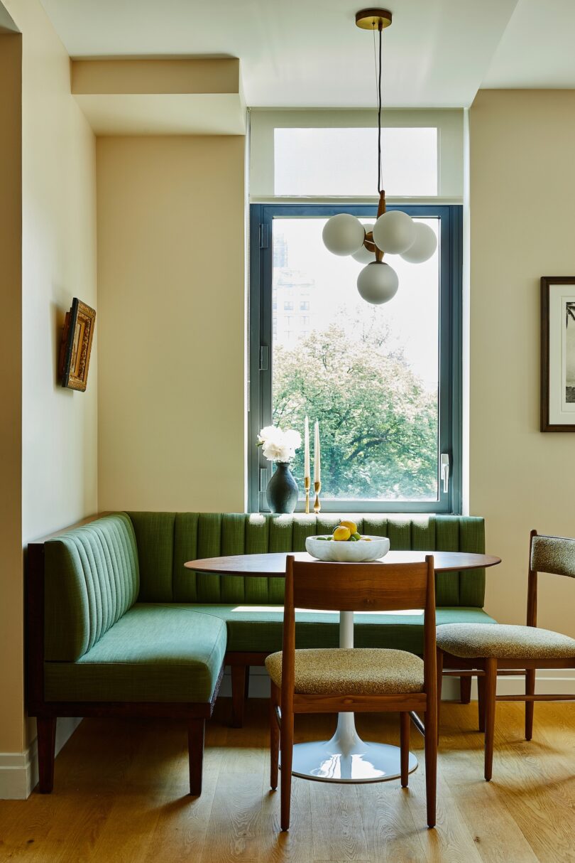 A small dining nook with a green upholstered bench, two chairs, a round table, a modern light fixture, and a window overlooking trees.