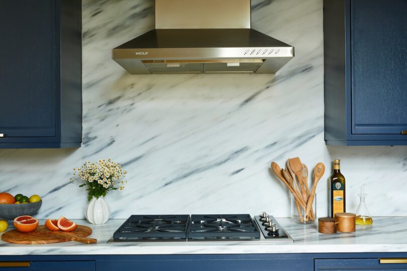 Modern kitchen with a marble backsplash, blue cabinets, a stainless steel range hood, gas stove, and countertop items including fruit, flowers, utensils, and a bottle of olive oil.
