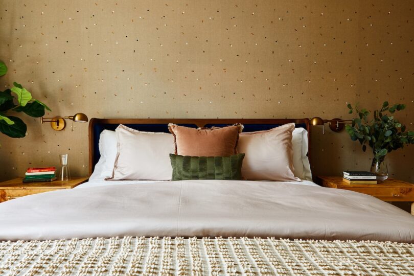A neatly made bed with pillows and a throw blanket, flanked by two wooden nightstands with books, a glass, and plants, set against a dotted beige wallpaper.