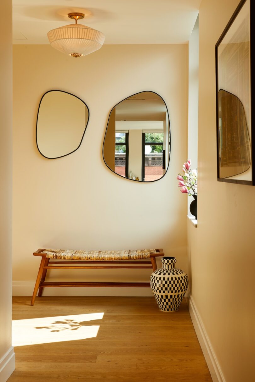 A hallway with two irregularly shaped mirrors, a woven bench, a checkered vase on the floor, a small window with pink flowers, and light wood flooring.
