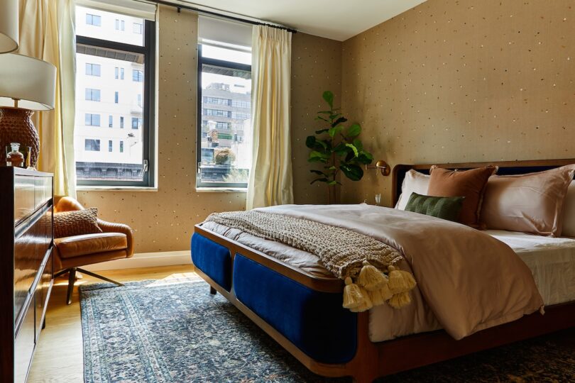 A modern bedroom with a neatly made bed, a patterned rug, two large windows with cream curtains, a green plant, and a brown leather chair by a wooden dresser.