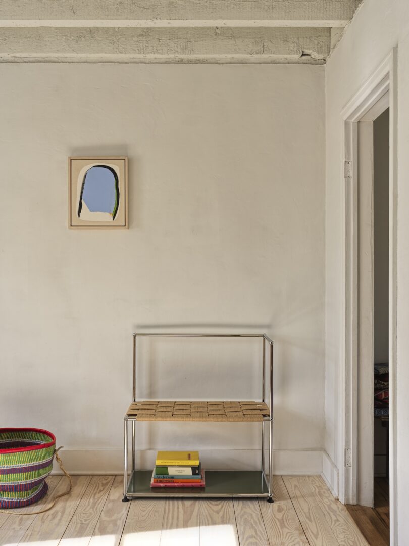 Minimalist interior with a wicker and metal bench, colorful books underneath, a multicolored basket on the floor, and abstract art on a white wall.