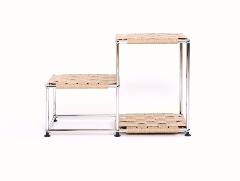 A two-tiered, minimalist shelving unit with metal frame and woven beige surfaces, arranged in a stepped configuration against a white background.
