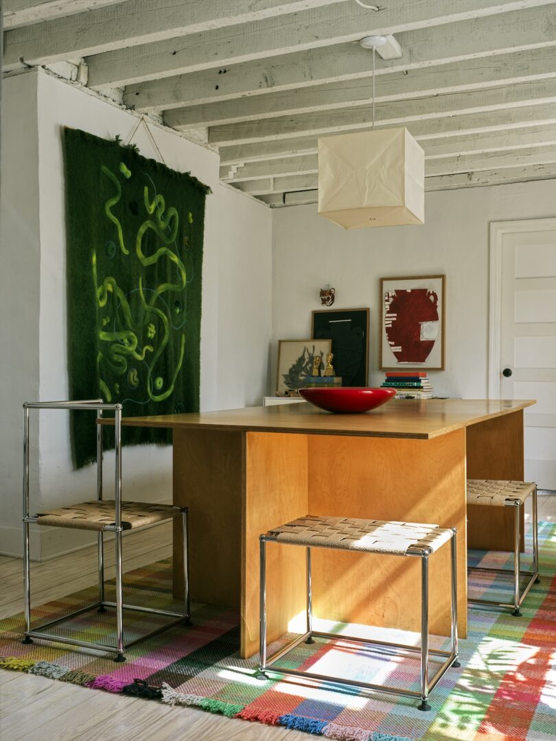 A modern room with a wooden table, metal-frame chairs with woven seats, colorful rug, abstract wall art, and a cube-shaped ceiling light.