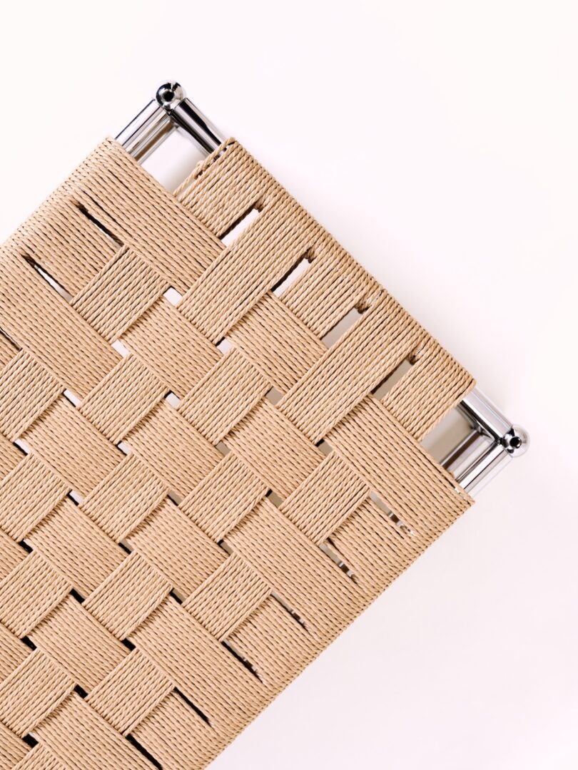 Close-up of a woven beige fabric strap pattern on a metal frame, photographed against a light background.