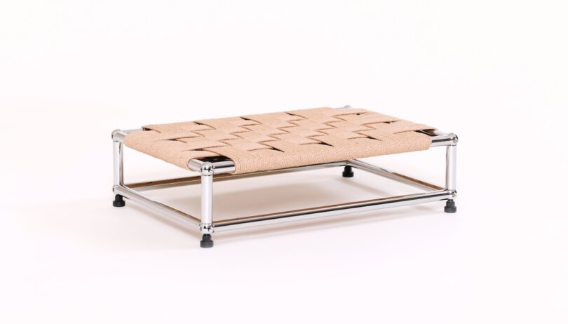 A low, rectangular platform with a metal frame and a woven, tan-colored surface, raised slightly above the ground on black feet.