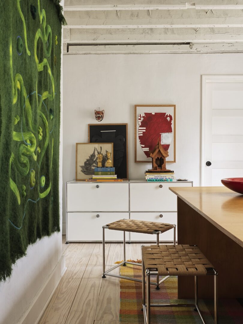 A modern room with a green abstract wall hanging, two white dressers topped with art and books, woven stools, a wooden table, and exposed beam ceiling.