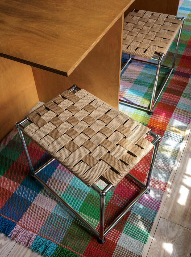 Two woven strap stools with metal frames are placed under a wooden table on a colorful checkered rug.