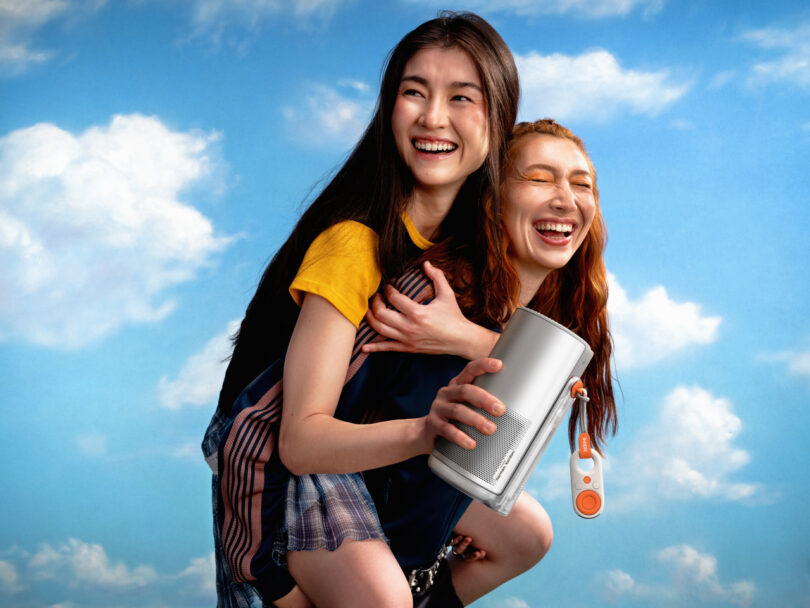 Two young women smiling, one giving the other a piggyback ride. The woman in front holds an XGIMI portable speaker with a strap, standing against a blue sky with clouds.