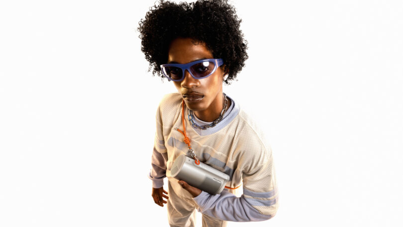 Person wearing a white outfit and blue sunglasses holds a compact XGIMI portable speaker, with a neutral expression, against a plain white background.