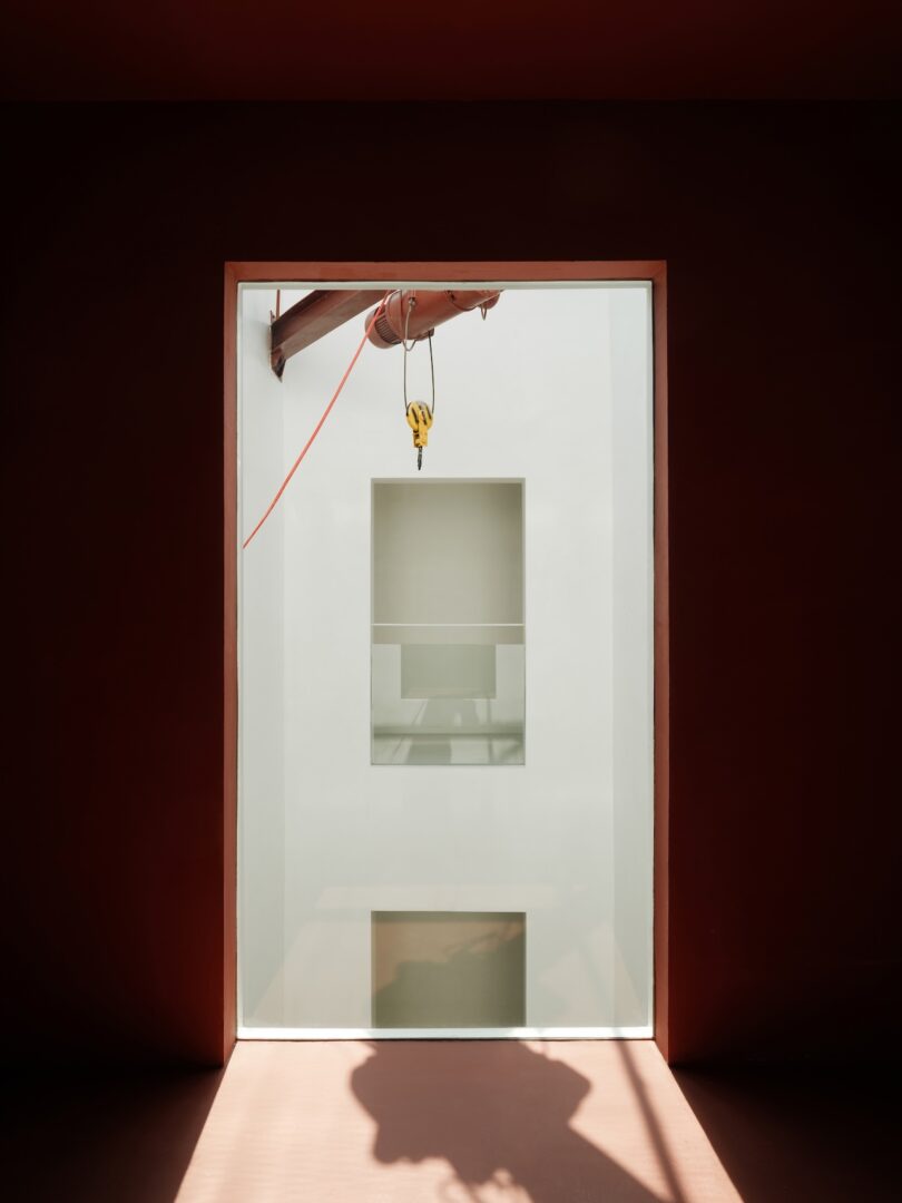 A large window frames a view of a white interior wall with geometric cut-outs; an orange construction hoist and cable are visible at the top.