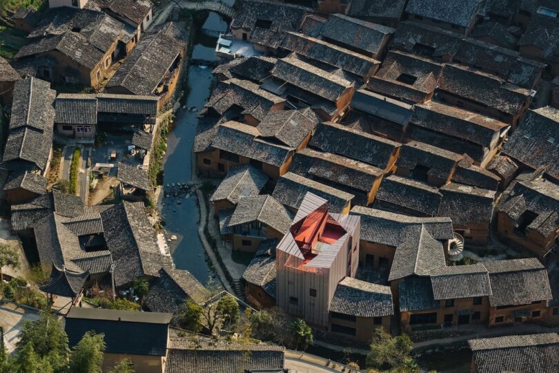 Aerial view of a traditional village with dark-tiled roofs and narrow canal, featuring one modern, light-colored building standing out among the older structures.
