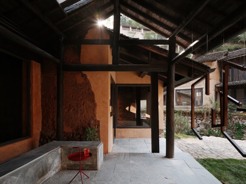 Courtyard of a rustic house with mud walls, slate floor, wooden beams, a built-in stone bench, a small red table, and an open window facing a garden area.