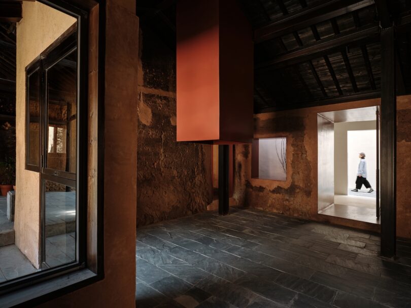 A person walks through a bright doorway in a rustic room with textured walls, slate floor, large windows, and a suspended red structure.