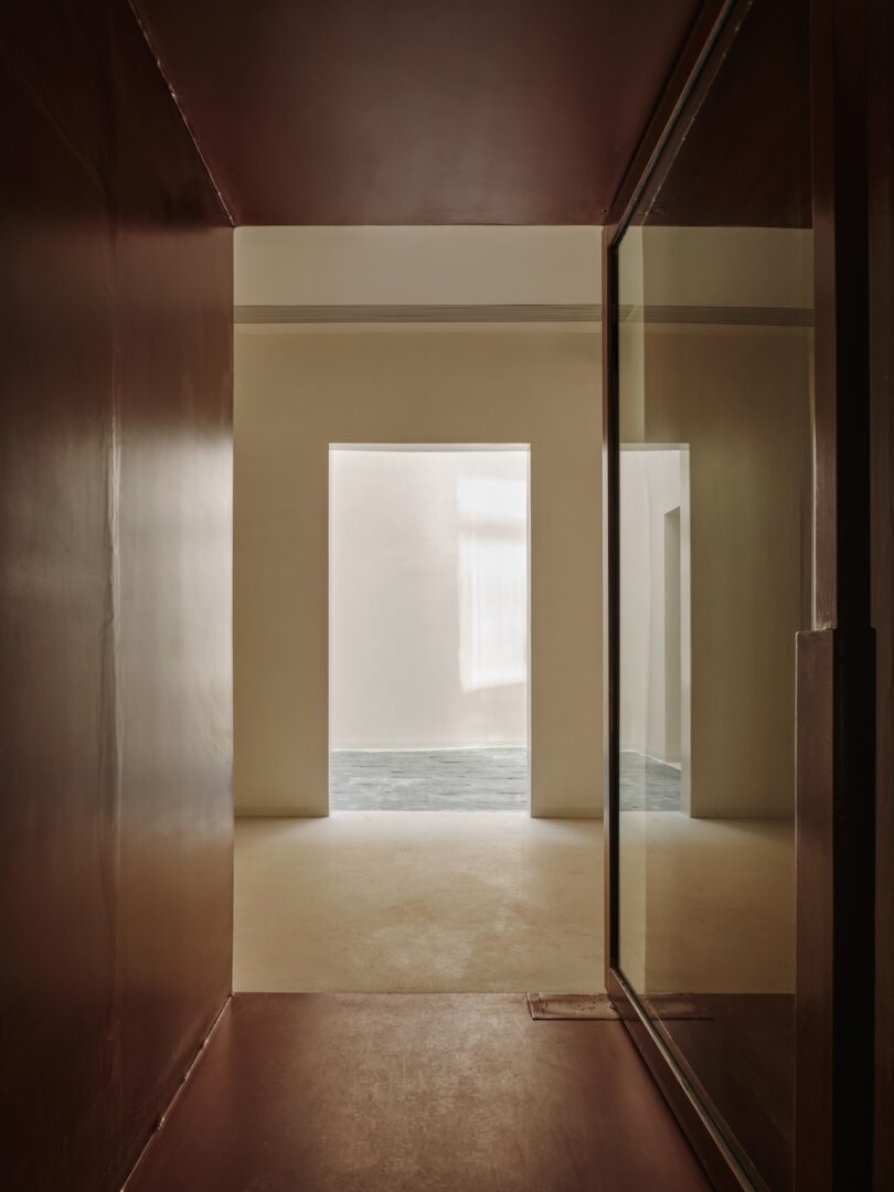 A minimalist hallway with brown walls, a reflective glass panel, and an open doorway leading to a bright room with cream-colored walls and a gray floor.