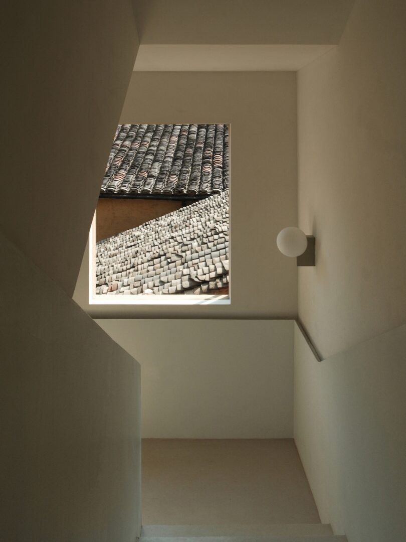 A view of tiled rooftops through a rectangular window at the end of a minimalist beige staircase with a round wall light.