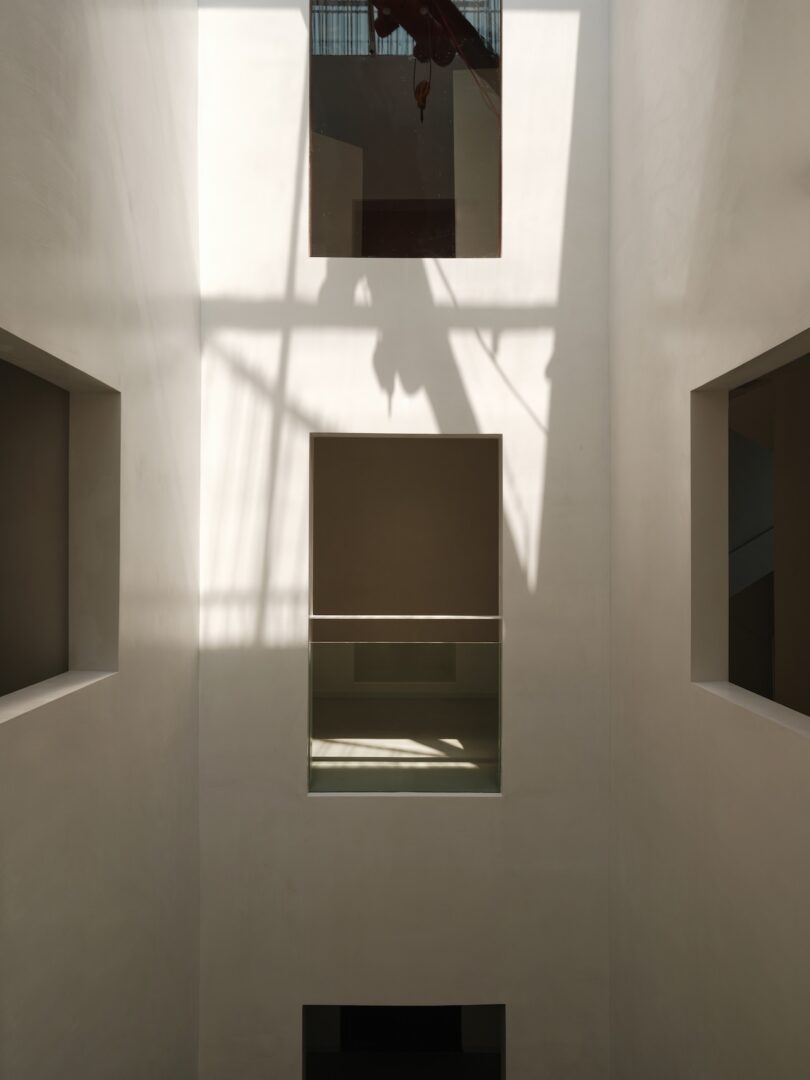 Tall white-walled interior with rectangular cutouts and natural light casting shadows through a grid-like window above.