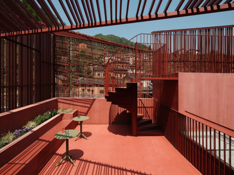 A rooftop terrace with red flooring, red metal railings, a spiral staircase, two small round tables, and views of a hillside town in the background.