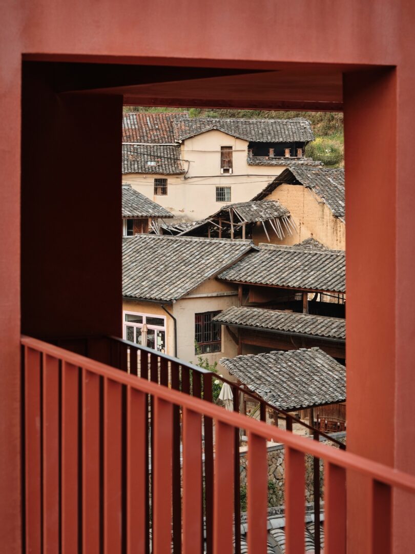 View through a red rectangular frame and railing, showing traditional buildings with tiled roofs in the background.
