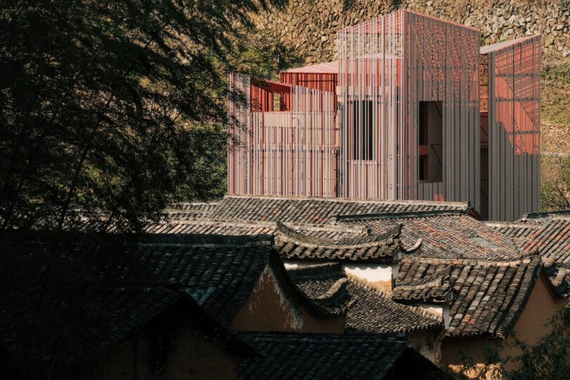 A modern pink and red rectangular building with vertical slats rises above traditional dark-tiled rooftops surrounded by trees and a stone wall.