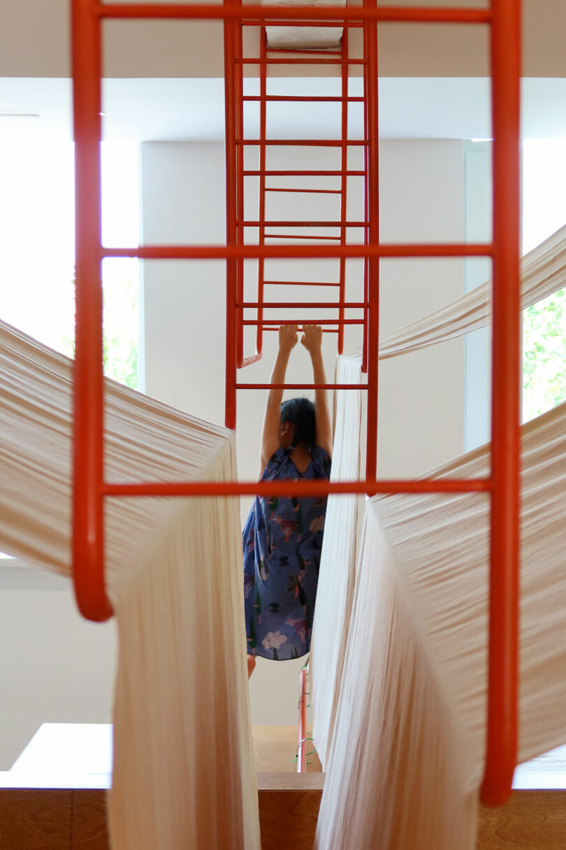 A person in a blue dress hangs from a red metal ladder structure, surrounded by draped white fabric in a brightly lit room.