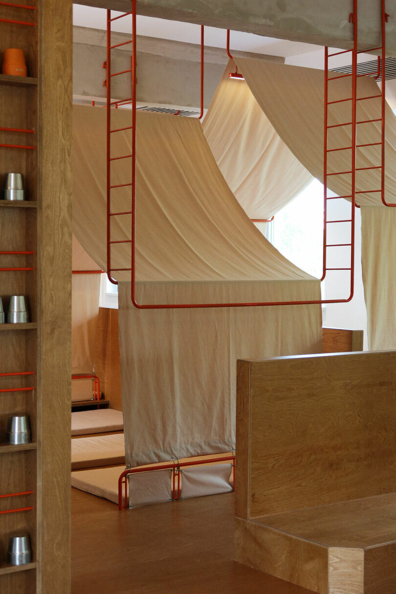 A modern interior with wooden furniture, metal ladders, hanging white fabric canopies, and neatly stacked metal cups on shelves.