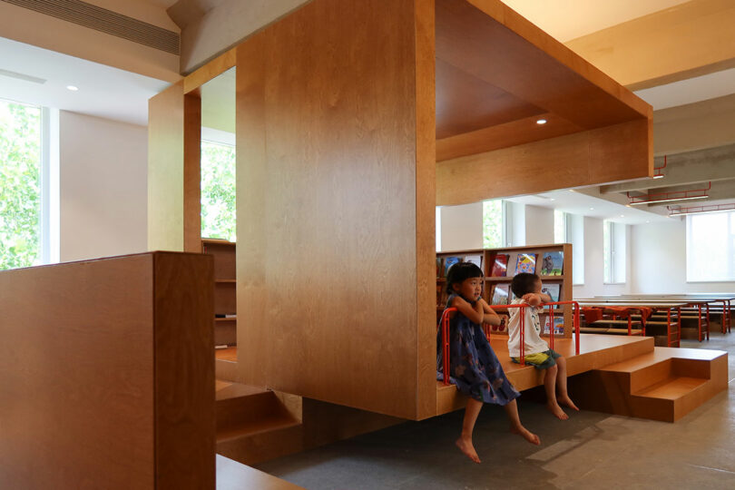 Two children sit on red bars attached to a modern wooden play structure inside a bright room with bookshelves and large windows.