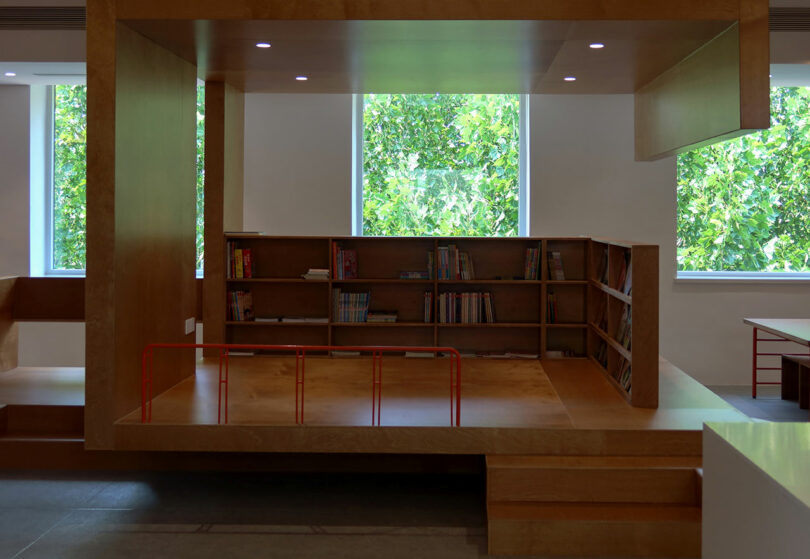 A modern reading nook with built-in wooden bookshelves, a raised platform, and large windows showing green foliage outside.