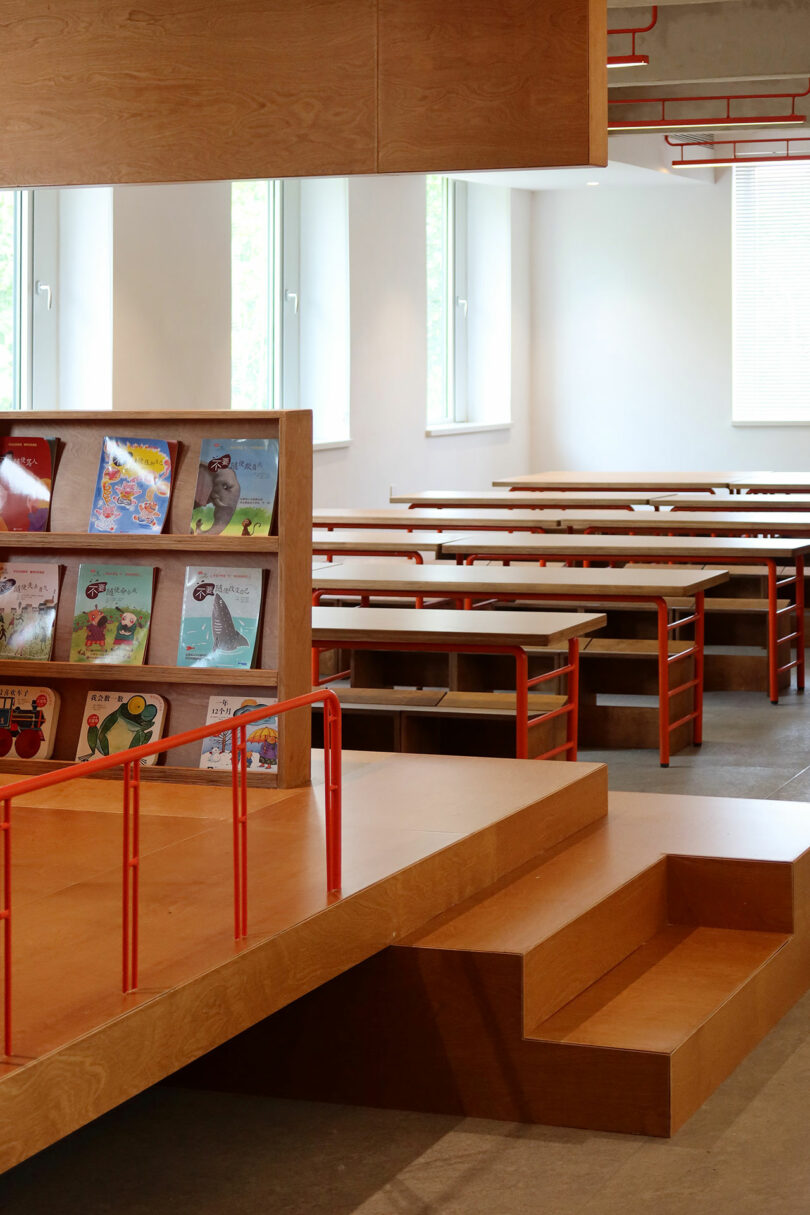 A modern room with wooden benches and tables, a stepped platform, and a bookshelf displaying children's books with orange railings. Large windows provide natural light.